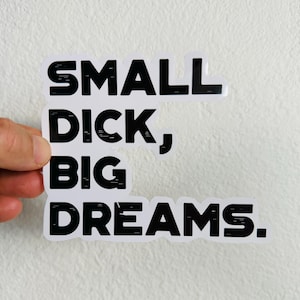 May include: A white sticker with black text that reads "SMALL DICK, BIG DREAMS." The sticker is held by a hand against a white background. The text is in a bold, sans-serif font.