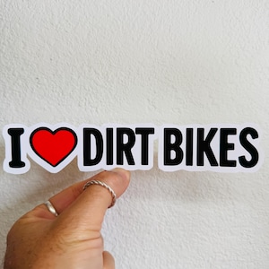Puede incluir: Una pegatina blanca con texto negro que dice "I ❤️ DIRT BIKES". El símbolo del corazón es rojo con un contorno negro. La pegatina está sujeta por una mano sobre un fondo blanco.