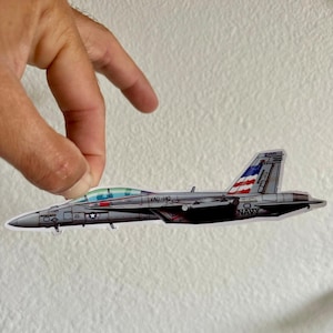 May include: A grey jet sticker with the text "NAVY" and an American flag design on the tail. The sticker also has the text "VAG-140". The jet has a green tinted cockpit.