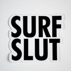 May include: A white sticker with the words "SURF SLUT" in bold, black capital letters. The text is centered and has a slight drop shadow effect, creating a 3D appearance. The sticker is rectangular with rounded corners.