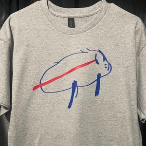 May include: A gray t-shirt featuring a blue and red graphic design. The design appears to be a stylized animal or creature with a red stripe across it. The shirt is made of a soft material and has a crew neck.