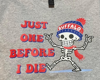 May include: A heather gray t-shirt featuring a skeleton wearing a blue and red Buffalo beanie and a red scarf. The shirt has the text "JUST ONE BEFORE I DIE" in red and blue lettering.