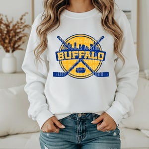May include: White sweatshirt featuring a blue and yellow Buffalo hockey-themed graphic. The design includes crossed hockey sticks, a puck, and the word "BUFFALO" in bold letters. Additional text reads "Prints Anything" and "Prints Anything".