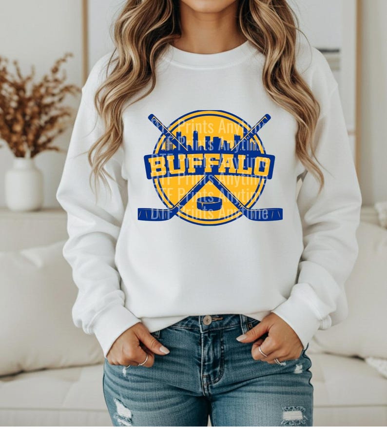 May include: White sweatshirt featuring a blue and yellow Buffalo hockey-themed graphic. The design includes crossed hockey sticks, a puck, and the word "BUFFALO" in bold letters. Additional text reads "Prints Anything" and "Prints Anything".