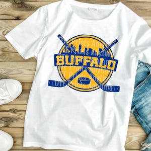 May include: White t-shirt featuring a yellow and blue graphic with the word "BUFFALO" in a blue rectangle. The design includes crossed hockey sticks, a hockey puck, and a city skyline. The shirt is made of a soft material.
