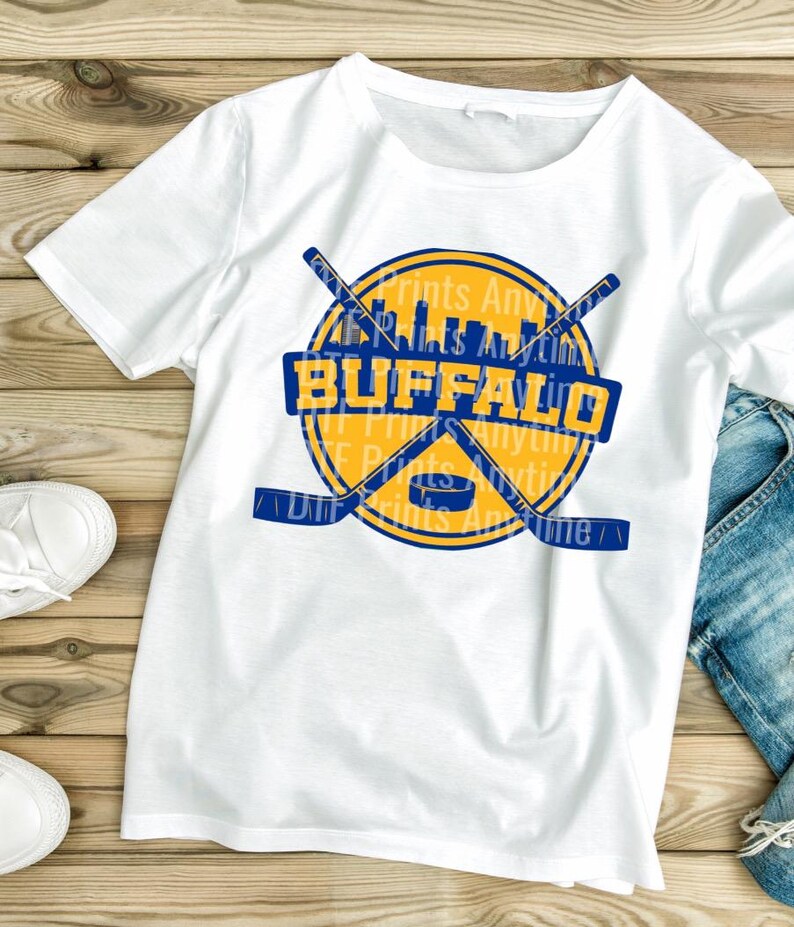 May include: White t-shirt featuring a yellow and blue graphic with the word "BUFFALO" in a blue rectangle. The design includes crossed hockey sticks, a hockey puck, and a city skyline. The shirt is made of a soft material.