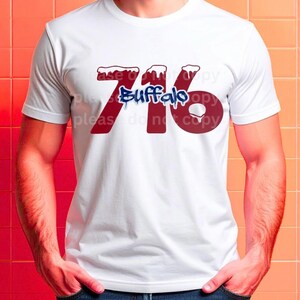 May include: White t-shirt featuring the text "716 Buffalo" in red and blue. The numbers and the word "Buffalo" are designed with a dripping effect, and the numbers have a snow-like design on top.