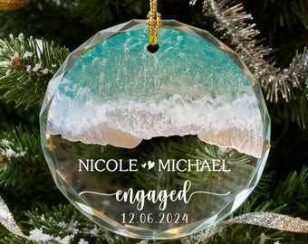 May include: A clear, round Christmas ornament with a beach scene design. The ornament has the names "Nicole" and "Michael" with the word "engaged" and the date "12.06.2024" printed on it. The ornament is hanging on a gold string.