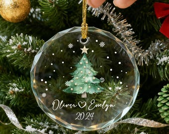 May include: A clear, round Christmas ornament with a gold string, featuring a green Christmas tree design and the names "Oliver & Evelyn" with the year "2024". The ornament is held in front of a decorated Christmas tree with red bows and gold ornaments.
