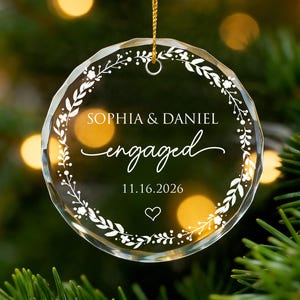 Może przedstawiać: Przezroczysta, okrągła szklana ozdoba choinkowa ze złotym sznurkiem. Ozdoba zawiera biały napis "SOPHIA & DANIEL engaged 11.16.2026", otoczony białym wieńcem kwiatowym i sercem. Świąteczna dekoracja.