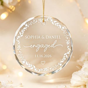 May include: A circular clear glass ornament featuring a white printed floral wreath design. The text reads SOPHIA & DANIEL, engaged, and 11.16.2026, with a small heart icon below the date. The ornament hangs from a thin gold cord.