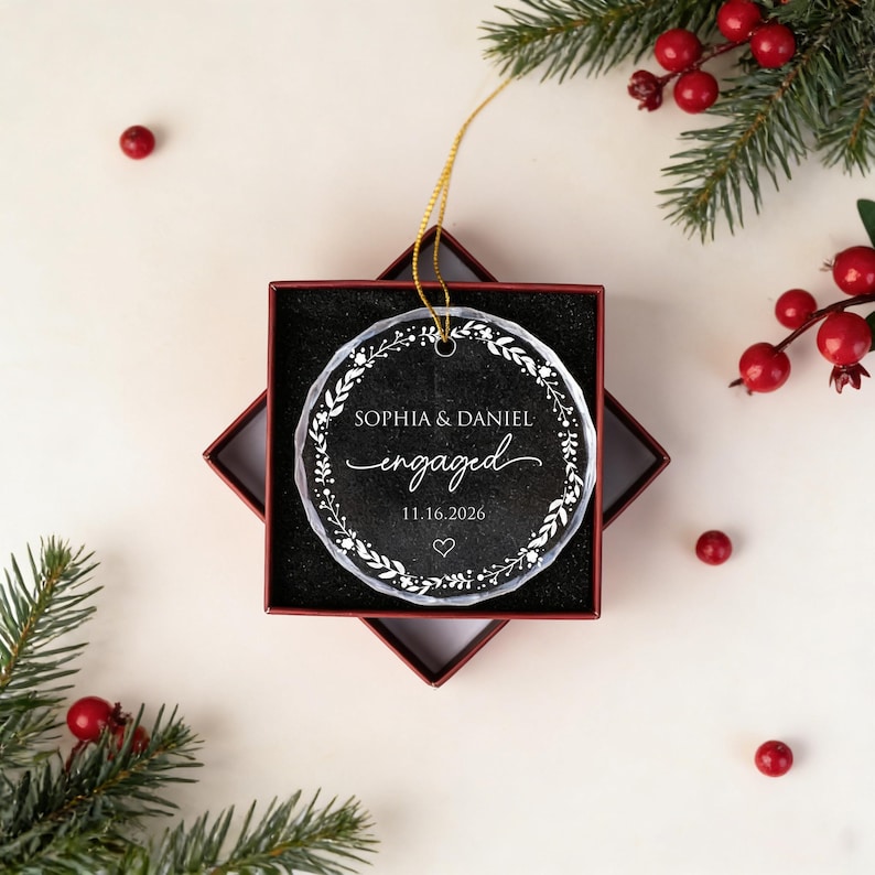 May include: A round, clear glass ornament with a scalloped edge, featuring a white printed wreath design, the text "SOPHIA & DANIEL", "engaged", and the date "11.16.2026" with a small heart, suspended by a gold string inside a square red box.