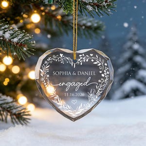 May include: A clear, faceted heart-shaped glass ornament hanging from a gold cord. The surface features white etched text reading SOPHIA & DANIEL, engaged, and 11.16.2026, surrounded by a delicate white floral wreath design and a small heart icon.