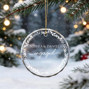 May include: A circular, faceted clear glass ornament hanging from a gold string. It features a white printed wreath design, the names SOPHIA & DANIEL, the word engaged in script, the date 11.16.2026, and a small heart outline at the bottom.