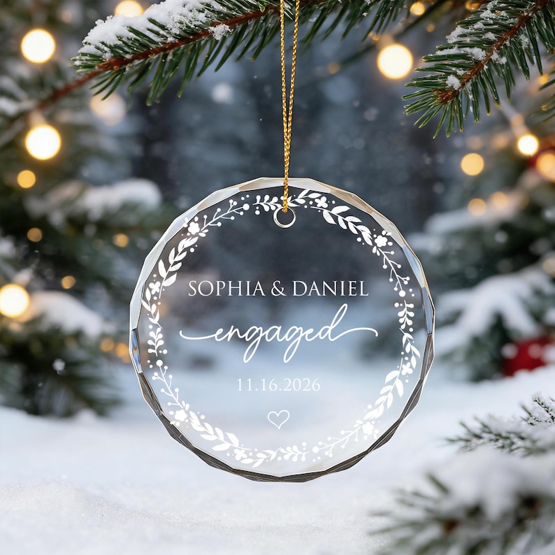 May include: A circular, faceted clear glass ornament hanging from a gold string. It features a white printed wreath design, the names SOPHIA & DANIEL, the word engaged in script, the date 11.16.2026, and a small heart outline at the bottom.