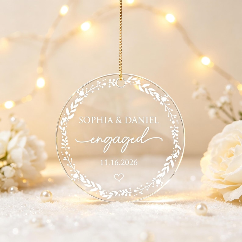 May include: A circular clear acrylic ornament hanging from a gold cord. The ornament features a white printed floral wreath design surrounding the text SOPHIA & DANIEL, the word engaged in script, the date 11.16.2026, and a small heart icon.