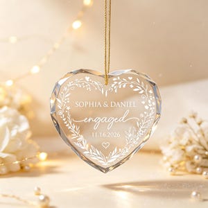 May include: A faceted clear glass heart ornament hanging from a gold cord. The center is etched with a white floral wreath design, the names SOPHIA & DANIEL, the word engaged, and the date 11.16.2026, finished with a small heart icon below.