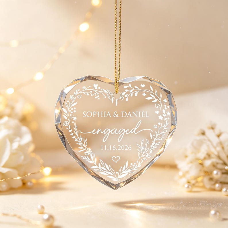 May include: A faceted clear glass heart ornament hanging from a gold cord. The center is etched with a white floral wreath design, the names SOPHIA & DANIEL, the word engaged, and the date 11.16.2026, finished with a small heart icon below.