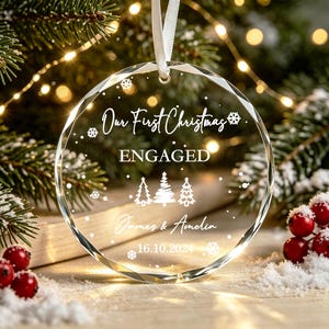 May include: A clear, circular, faceted glass ornament hanging from a white ribbon. The ornament features white etched text reading "Our First Christmas ENGAGED", "James & Amelia", and "16.10.2024", along with three small pine tree icons and snowflakes.