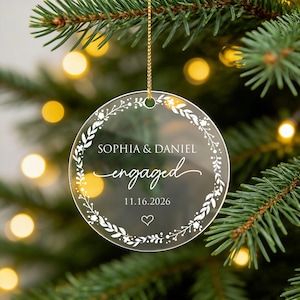 May include: A circular clear acrylic ornament featuring a white printed floral wreath design. The text reads SOPHIA & DANIEL, engaged, and 11.16.2026, with a small heart icon below. The ornament hangs from a thin gold string.