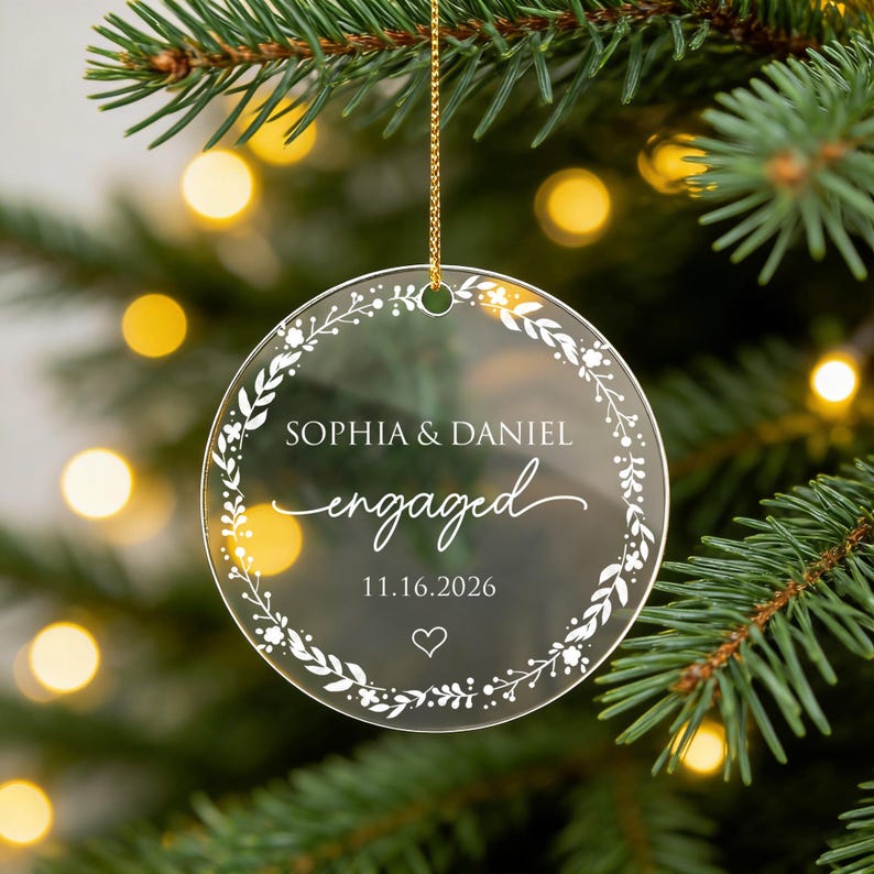 May include: A circular clear acrylic ornament featuring a white printed floral wreath design. The text reads SOPHIA & DANIEL, engaged, and 11.16.2026, with a small heart icon below. The ornament hangs from a thin gold string.