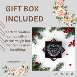 May include: A clear, heart-shaped glass ornament featuring the engraved text "SOPHIA & DANIEL engaged 11.16.2026" surrounded by a leaf wreath. The ornament hangs by a gold string inside a square red gift box, surrounded by pine branches and red berries.