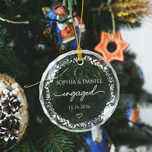 May include: A circular, faceted clear acrylic ornament hanging by a gold string. The ornament features a white printed floral wreath design surrounding the text "SOPHIA & DANIEL", "engaged", the date "11.16.2026", and a small heart icon.