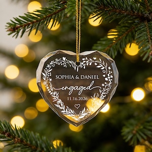 May include: A faceted heart-shaped clear glass ornament hanging from a gold string. The center features white etched text reading SOPHIA & DANIEL, engaged, and 11.16.2026, surrounded by a delicate white floral wreath design with a small heart at the bottom.