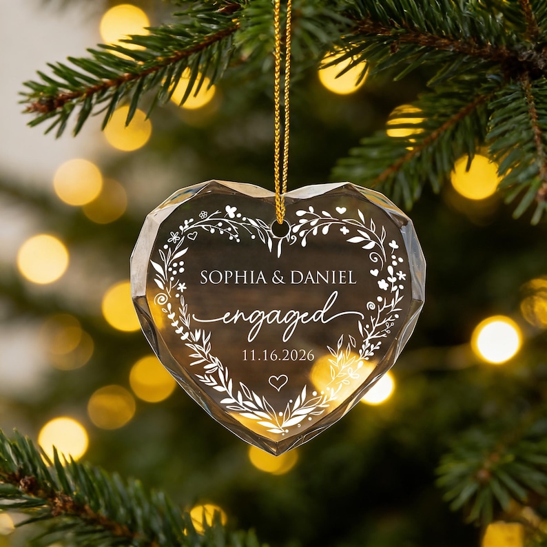 May include: A faceted heart-shaped clear glass ornament hanging from a gold string. The center features white etched text reading SOPHIA & DANIEL, engaged, and 11.16.2026, surrounded by a delicate white floral wreath design with a small heart at the bottom.