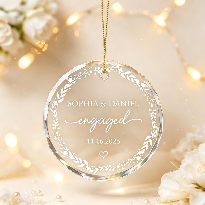 May include: A circular clear glass ornament featuring a white printed floral wreath design. The text reads SOPHIA & DANIEL, engaged, and 11.16.2026, with a small heart icon below the date. The ornament hangs from a thin gold cord.