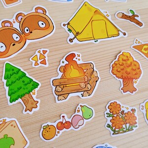 Op de afbeelding: Een verzameling kleurrijke stickers met cartoon illustraties van kampeer- en natuurelementen. Inclusief een gele tent, kampvuur, bomen, fruit en dierenfiguren. De stickers zijn gerangschikt op een licht houten oppervlak.