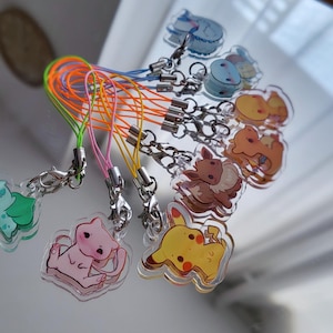 Op de afbeelding: Een set van negen kleurrijke sleutelhangers met verschillende cartoonfiguren. Elke sleutelhanger heeft een heldere acrylbedel met een ander figuurontwerp en een kleurrijk koord dat is bevestigd aan een zilveren metalen sluiting.