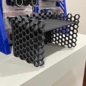 May include: A black, 3D-printed shelf with a honeycomb design. The shelf is designed to hold toy cars, with a flat surface and sides made of interconnected hexagonal shapes. The shelf is displayed on a white surface.