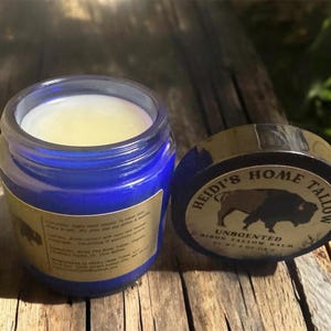 May include: A blue glass jar of unscented bison tallow balm with the lid off, next to the lid. The lid is black with gold lettering and a bison graphic. The jar has a gold label with text.