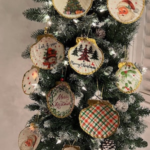 May include: A Christmas tree decorated with various ornaments made from seashells. The ornaments feature festive designs, including a snowman, a Christmas tree, Santa Claus, and the phrase "Merry Christmas".
