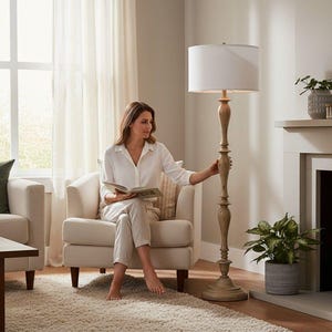 May include: A tall floor lamp featuring a light brown, turned wood-style base with a circular pedestal and a simple, cylindrical white fabric lampshade. The lamp stands upright on a floor, providing illumination through its white shade.