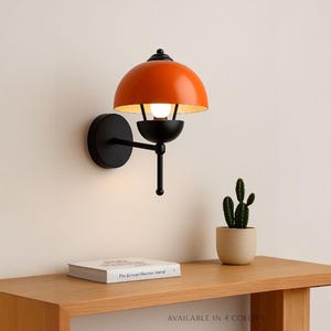 Puede incluir: Lámpara de pared naranja y negra con una pantalla en forma de cúpula. La lámpara tiene un brazo y una base negros, y una bombilla es visible dentro de la pantalla. Un pequeño cactus en una maceta beige está sobre una mesa de madera.