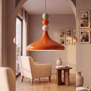 Op de afbeelding: Een hanglamp met een grote, glanzende oranje klokvormige kap die aan een zwart snoer hangt. Boven de kap bevat het armatuur drie gestapelde bolvormige accenten in de kleuren lichtgroen, feloranje en wit.