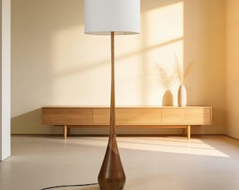 May include: A floor lamp featuring a tall, slender wooden stem that transitions into a wider, teardrop-shaped base made of dark wood. The lamp is topped with a large, cylindrical white fabric lampshade and includes a black power cord.