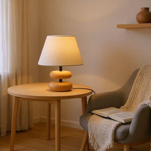 Pode incluir: Uma luminária de mesa de madeira com uma cúpula bege e uma base de madeira empilhada. A luminária está em uma mesa lateral redonda de madeira. Uma poltrona cinza com um cobertor creme e um livro aberto estão por perto.