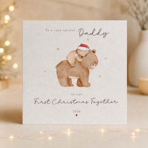May include: A square greeting card featuring a watercolour illustration of a large brown bear carrying a small bear wearing a red Santa hat. The text reads: To a very special Daddy on our First Christmas Together 2026, accented with small gold stars.