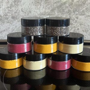 Pode incluir: Uma coleção de pequenos frascos transparentes com tampas pretas, cheios de vários pós e grânulos coloridos. Os frascos estão dispostos em forma de pirâmide, mostrando as diferentes cores e texturas dos conteúdos.