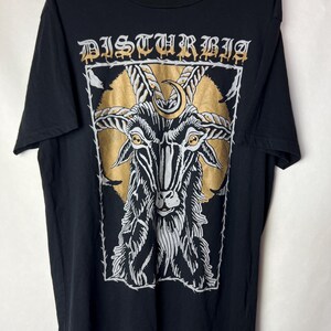 May include: Black t-shirt featuring a goat illustration with gold accents and the word "DISTURBIA" in a gothic font. The goat has large horns and a crescent moon. The design is framed by a white border.
