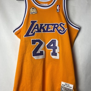 Könnte beinhalten: Ein goldgelbes und lila Lakers Basketballtrikot mit der Nummer 24 auf der Vorderseite. Das Trikot hat den Schriftzug "Lakers" in Lila auf der Brust und einen "60" Aufnäher. Das Trikot hängt an einem schwarzen Kleiderbügel.