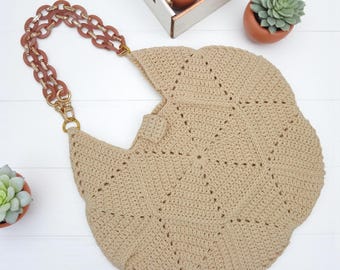 May include: A beige crochet bag featuring a geometric pattern and a thick, brown, interlocking plastic chain handle. The bag has a rounded, scalloped edge design and is constructed from a textured yarn material with gold-toned metal hardware.