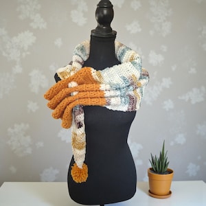 May include: A handmade crochet scarf and gloves set displayed on a black mannequin. The scarf features stripes of cream, blue, brown, and yellow. The gloves are a solid orange color. A small orange potted plant sits nearby.