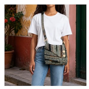 May include: A crossbody bag crafted from textured, multi-colored yarn in shades of green, brown, and black. The bag features a flap closure secured by two light-colored wooden buttons, a long shoulder strap, and circular wooden ring connectors.