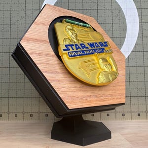 以下が含まれることがあります： 木製のフレームと黒いベースに収められた、金色のスターウォーズライバルラン10Kメダル。メダルにはキャラクターと「STAR WARS RIVAL RUN 10K 2019」の文字が描かれています。フレームは天然の木目仕上げです。
