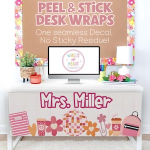 May include: A white desk with a decorative wrap featuring the text "Mrs. Miller" in pink, along with floral and school-themed graphics. The background includes a brown sign that reads "PEEL & STICK DESK WRAPS" and a computer monitor.
