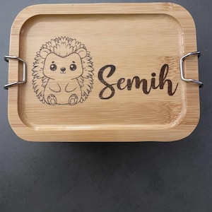 Könnte beinhalten: Eine Lunchbox aus Holz mit einem Klappdeckel und Metallverschlüssen. Die Lunchbox ist mit einem Comic-Igel verziert und der Name "Semih" ist auf der Vorderseite eingraviert.